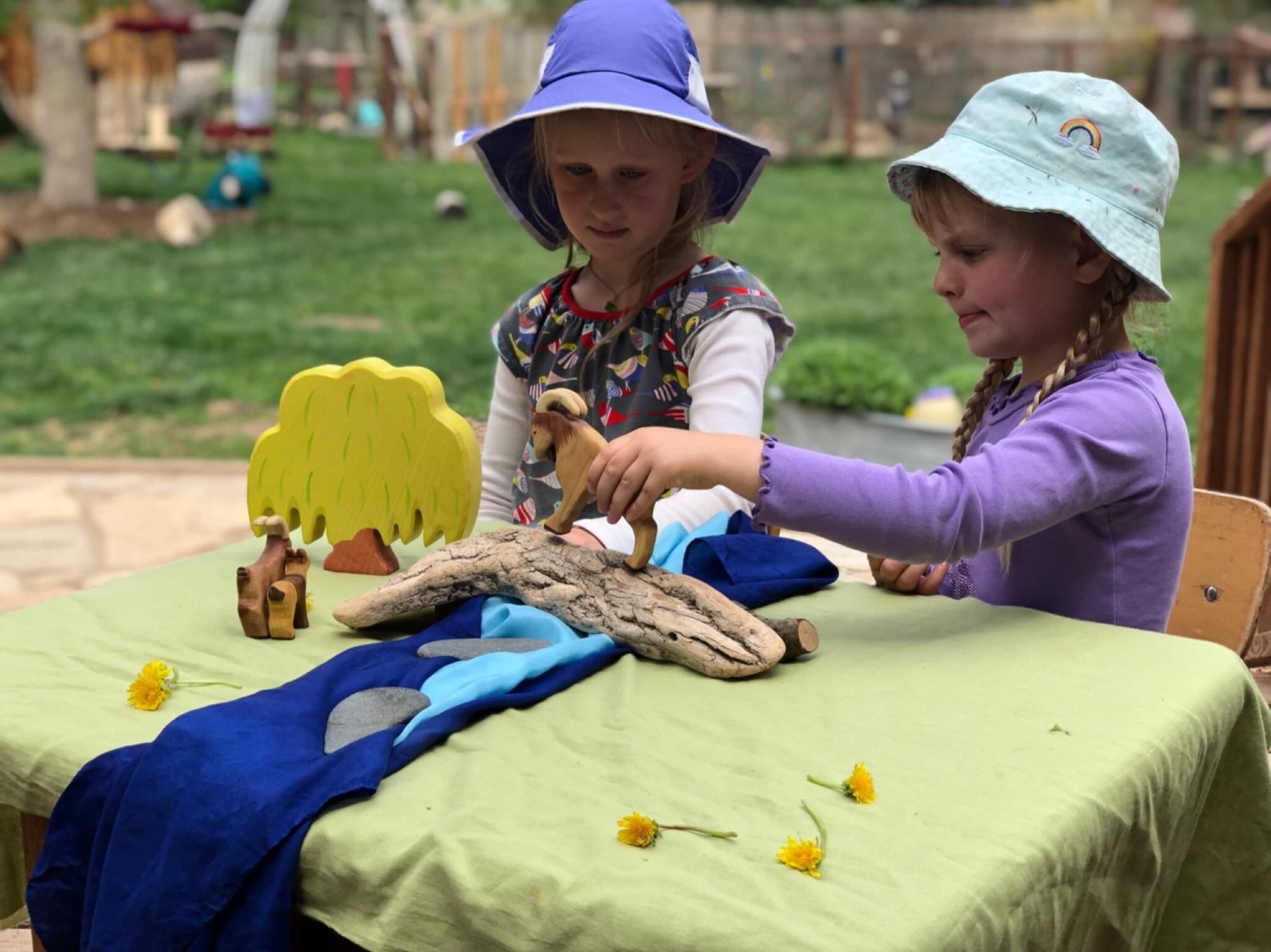 Two children playing with wooden animals and play silks at an outdoor table